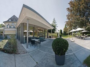 A modern outdoor seating area with tables and chairs, surrounded by a landscaped garden and a building with large windows.