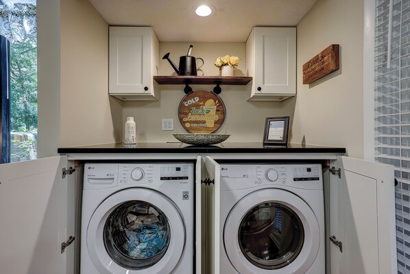 A laundry room with two front-loading washing machines, a shelf with a pitcher and flowers, and a sign on the wall. - Sunset Rentals | St. Andrews 1772 | Dog Friendly (Hilton Head)