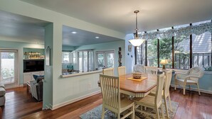 A spacious dining area with a wooden table and chairs, a television in the adjacent room, and large windows offering views of the outdoors. - Sunset Rentals | St. Andrews 1772 | Dog Friendly (Hilton Head)