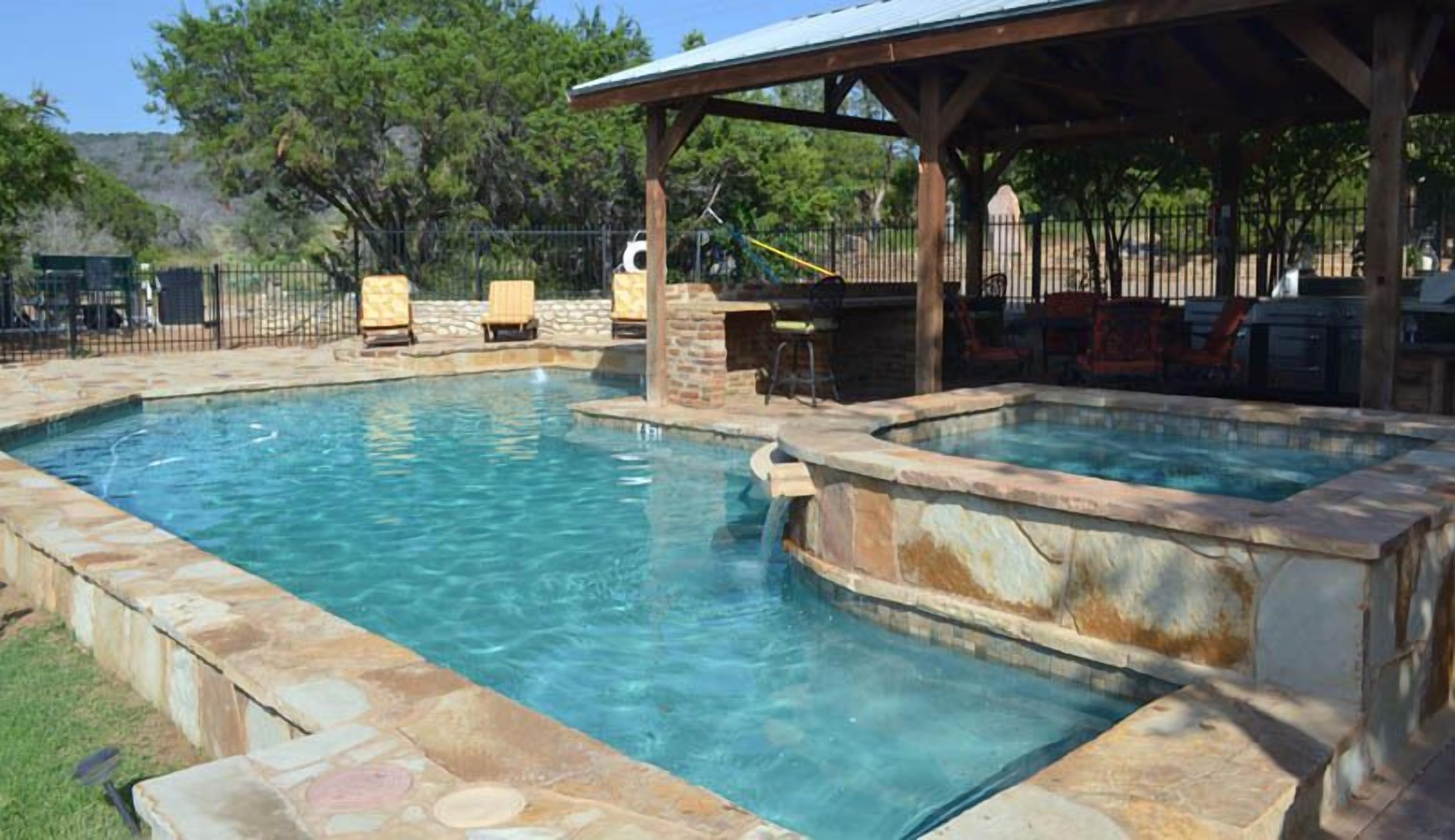 A swimming pool with a hot tub, stone borders, and a covered seating area with chairs.