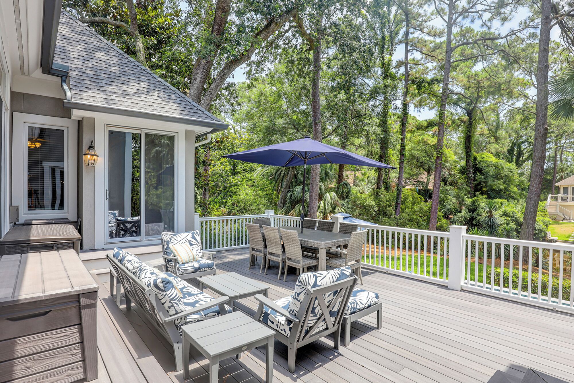A covered outdoor deck with a dining table and chairs, surrounded by a white railing and lush greenery.