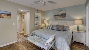 A bedroom with a bed, bedside tables, a ceiling fan, a bench, and a painting on the wall. - Sunset Rentals | St. Andrews 1876 | Dog Friendly (Hilton Head Island)