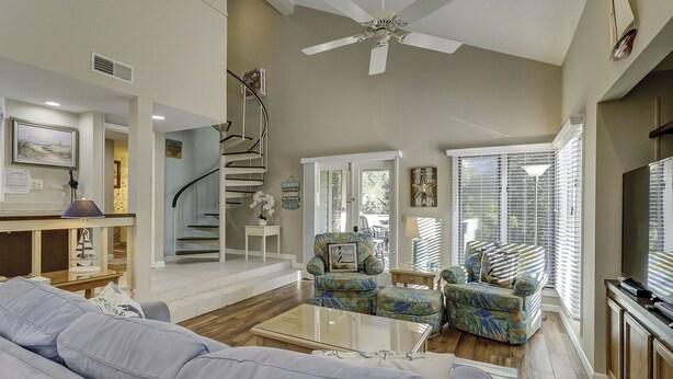 A spacious living room with a staircase, a ceiling fan, a television, and a view of the outdoors through large windows. - Sunset Rentals | St. Andrews 1876 | Dog Friendly (Hilton Head Island)