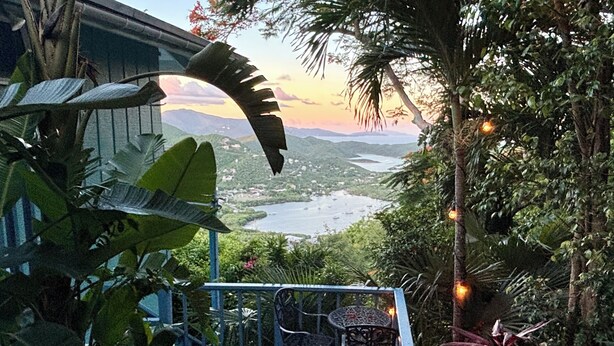 A terrace with chairs and a table, surrounded by lush greenery and a scenic view of a valley and water. - Beautiful Cottage w/Stunning View, Tesla Backup Power, Hot Tub, Family or Couple (St. John)