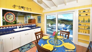 A bright yellow kitchen with a blue tiled backsplash, a large window with a view of the outdoors, and a dining area with yellow tableware. - Beautiful Cottage w/Stunning View, Tesla Backup Power, Hot Tub, Family or Couple (St. John)