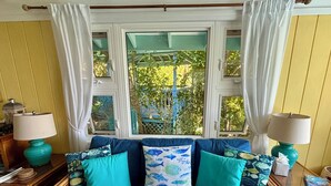 A room with a blue sofa, teal cushions, two lamps, and a view through a window with curtains. - Beautiful Cottage w/Stunning View, Tesla Backup Power, Hot Tub, Family or Couple (St. John)