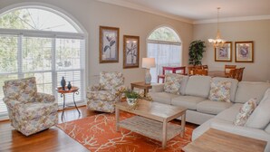 A spacious living room with a sofa, armchairs, and a coffee table. There are large windows with blinds, a chandelier, and framed artwork on the walls. - Sunset Rentals | Crabline 49 | Dog Friendly (Hilton Head Island)