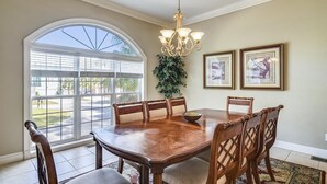 A dining room with a large table, six chairs, a chandelier, and two framed pictures on the wall. - Sunset Rentals | Crabline 49 | Dog Friendly (Hilton Head Island)