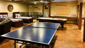 Ein Raum mit Tischtennisplatte, Billardtisch und Snookertisch. Zwei Sofas und ein Sessel stehen im Raum. Die Decke ist aus Holz und es gibt ein Fenster mit Jalousien.