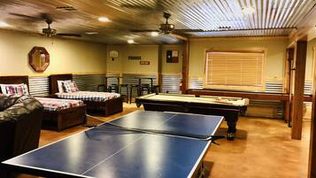 A room with a ping pong table, a pool table, and a billiards table. There are two couches and a chair. The room has a wooden ceiling and a window with blinds.