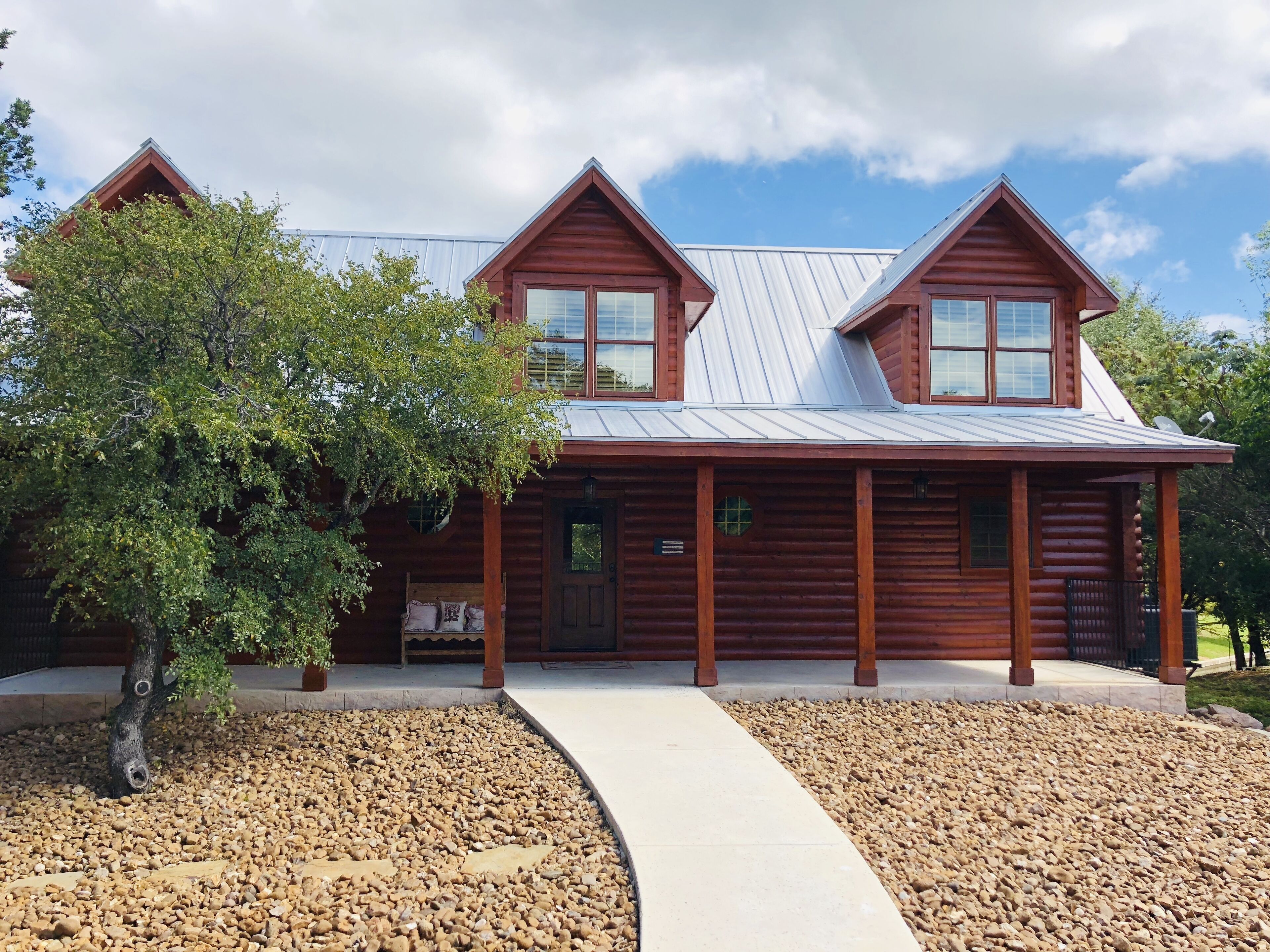 A log cabin with a metal roof, a porch, and a gravel driveway.