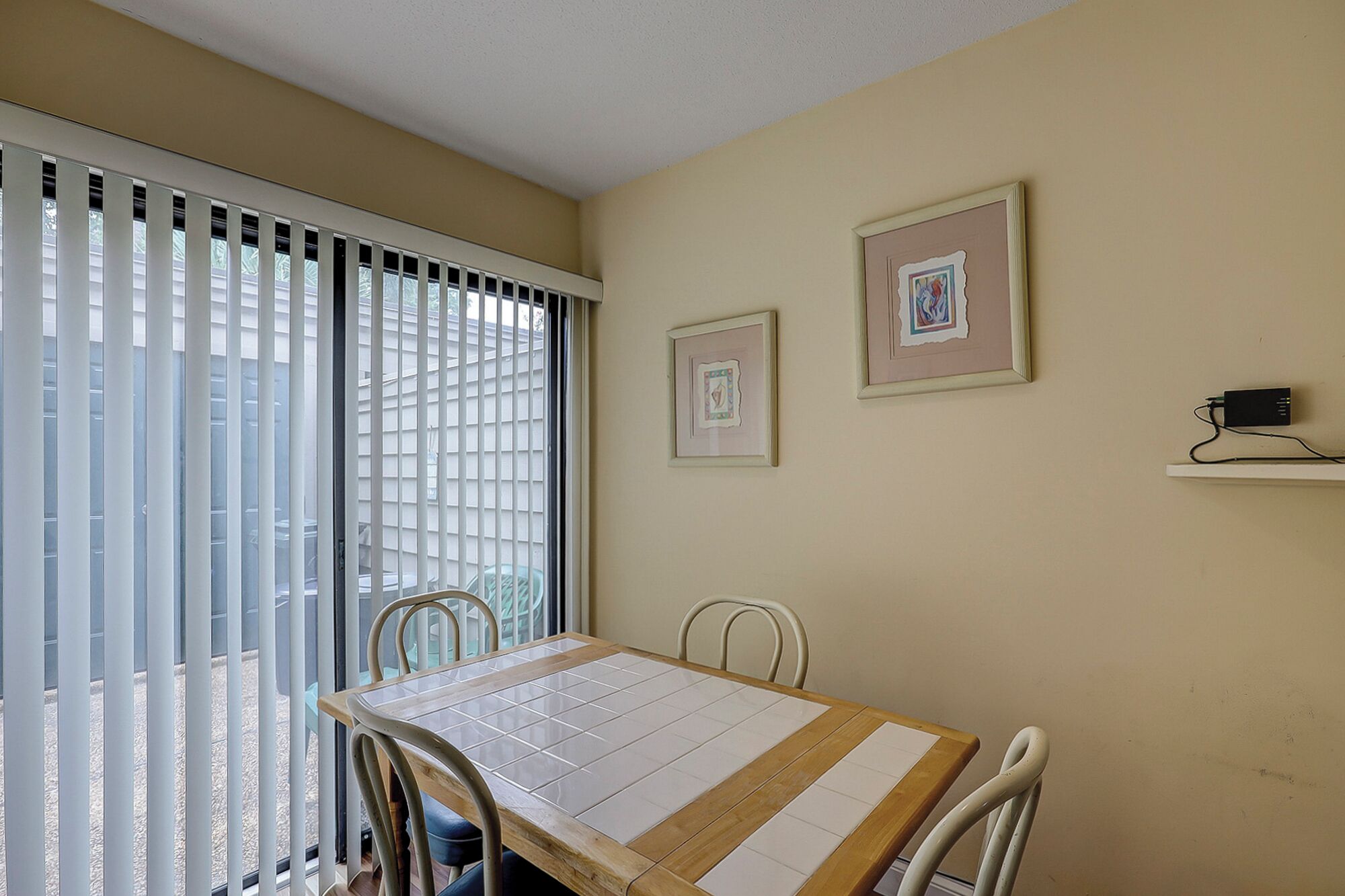 Un coin repas avec une table et des chaises, une fenêtre avec des stores et deux tableaux encadrés accrochés au mur.