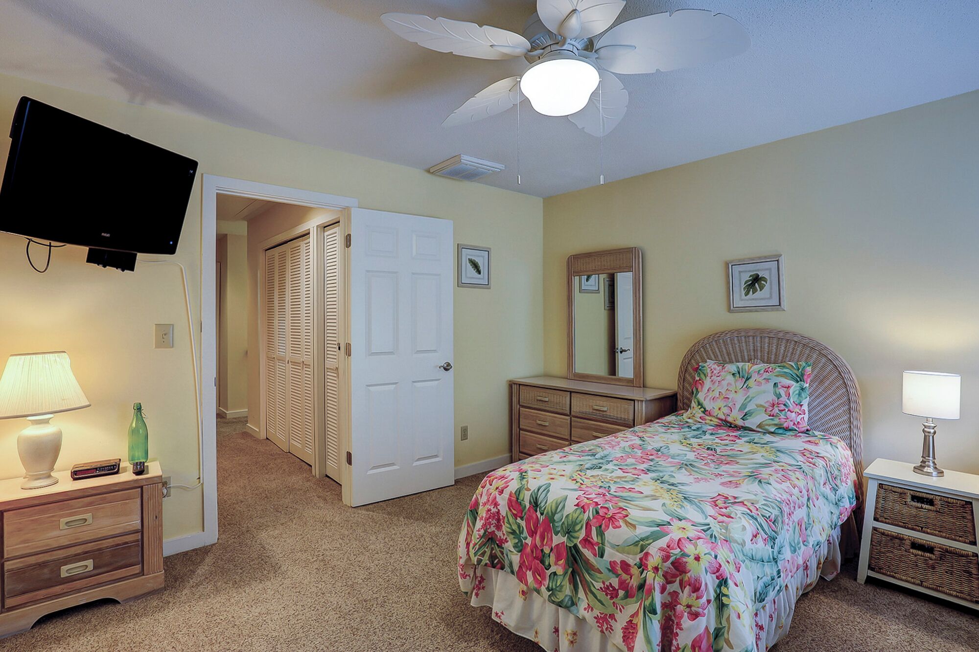 A bedroom with a bed, a dresser, a nightstand, a television, a ceiling fan, and a door leading to another room.