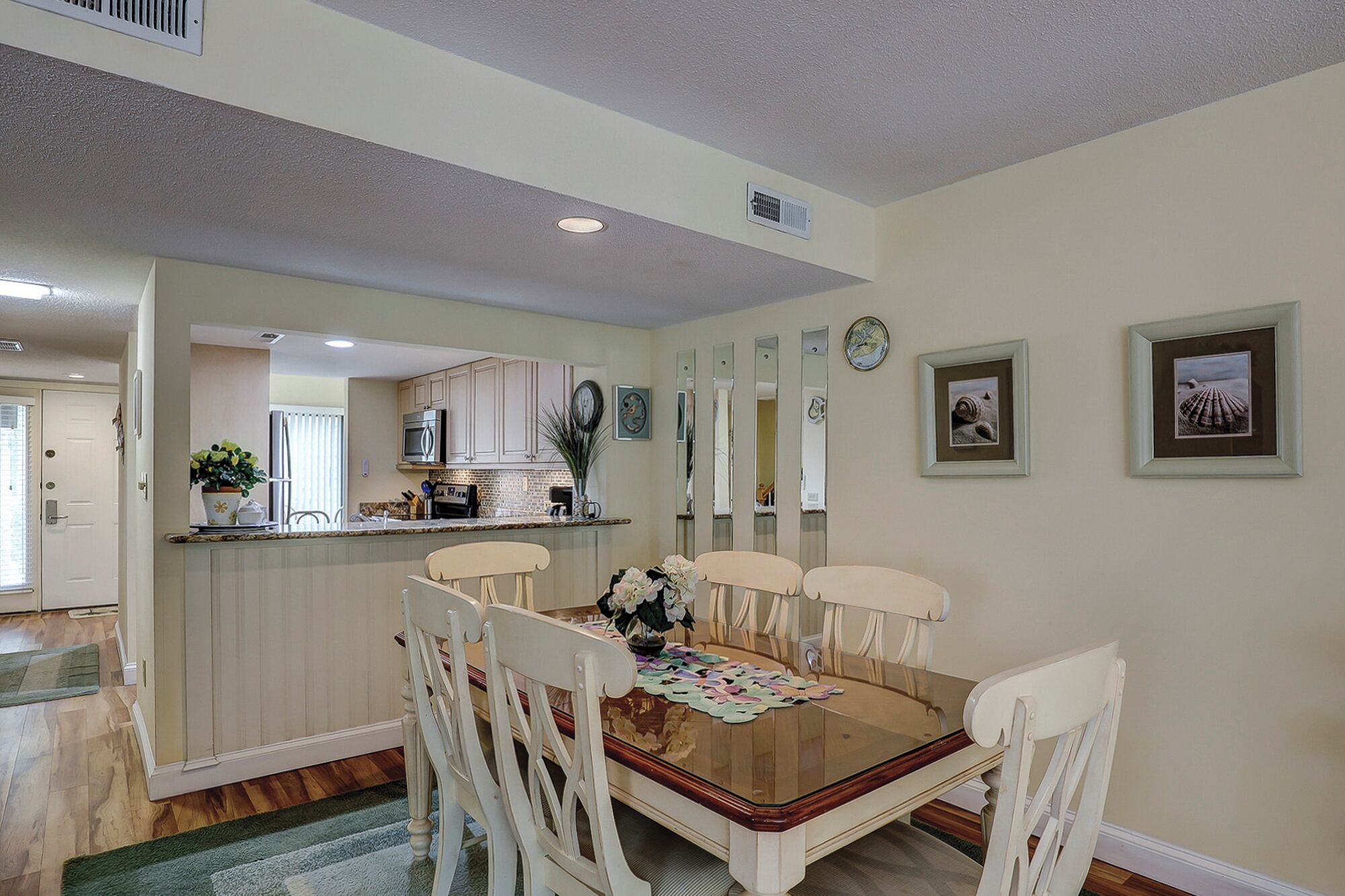A dining area with a wooden table and white chairs, a kitchen in the background, and a hallway leading to a door.