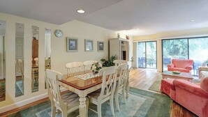 Un comedor con mesa de madera y sillas blancas, un sofá rojo y un amplio ventanal corredera que da a un patio con mobiliario exterior.