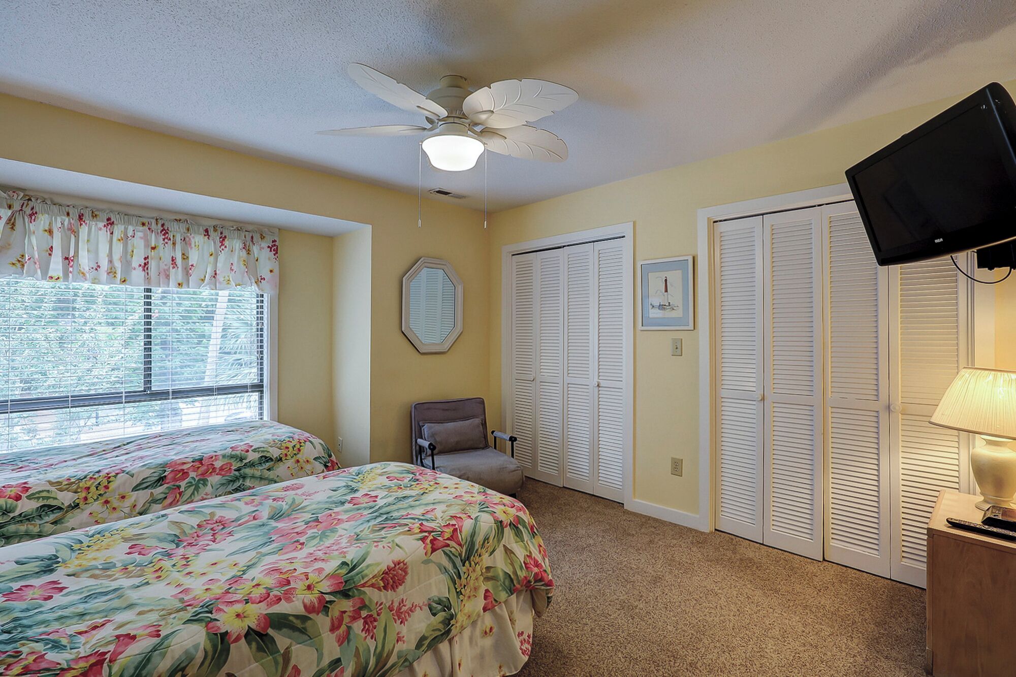 A bedroom with two beds, a ceiling fan, a window with blinds, a chair, and a TV on a stand.