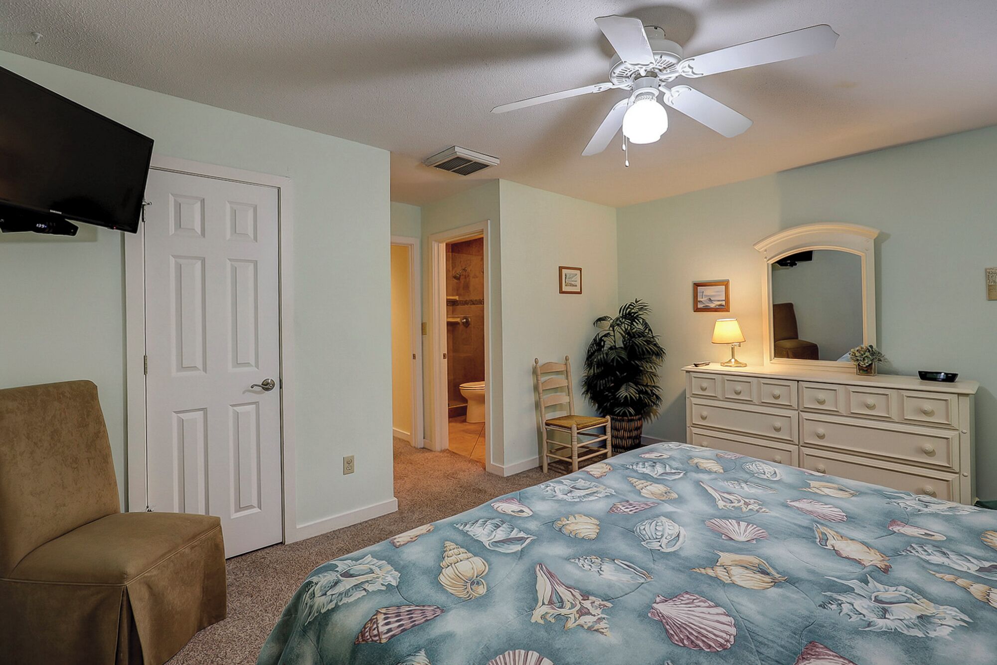 A bedroom with a bed, a dresser, a chair, a ceiling fan, and a door leading to a bathroom.