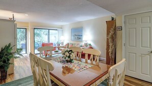 Un comedor con una mesa de madera, sillas y una mesa con cubierta de vidrio. Se observa una puerta a la derecha y una ventana con vistas a zonas verdes.