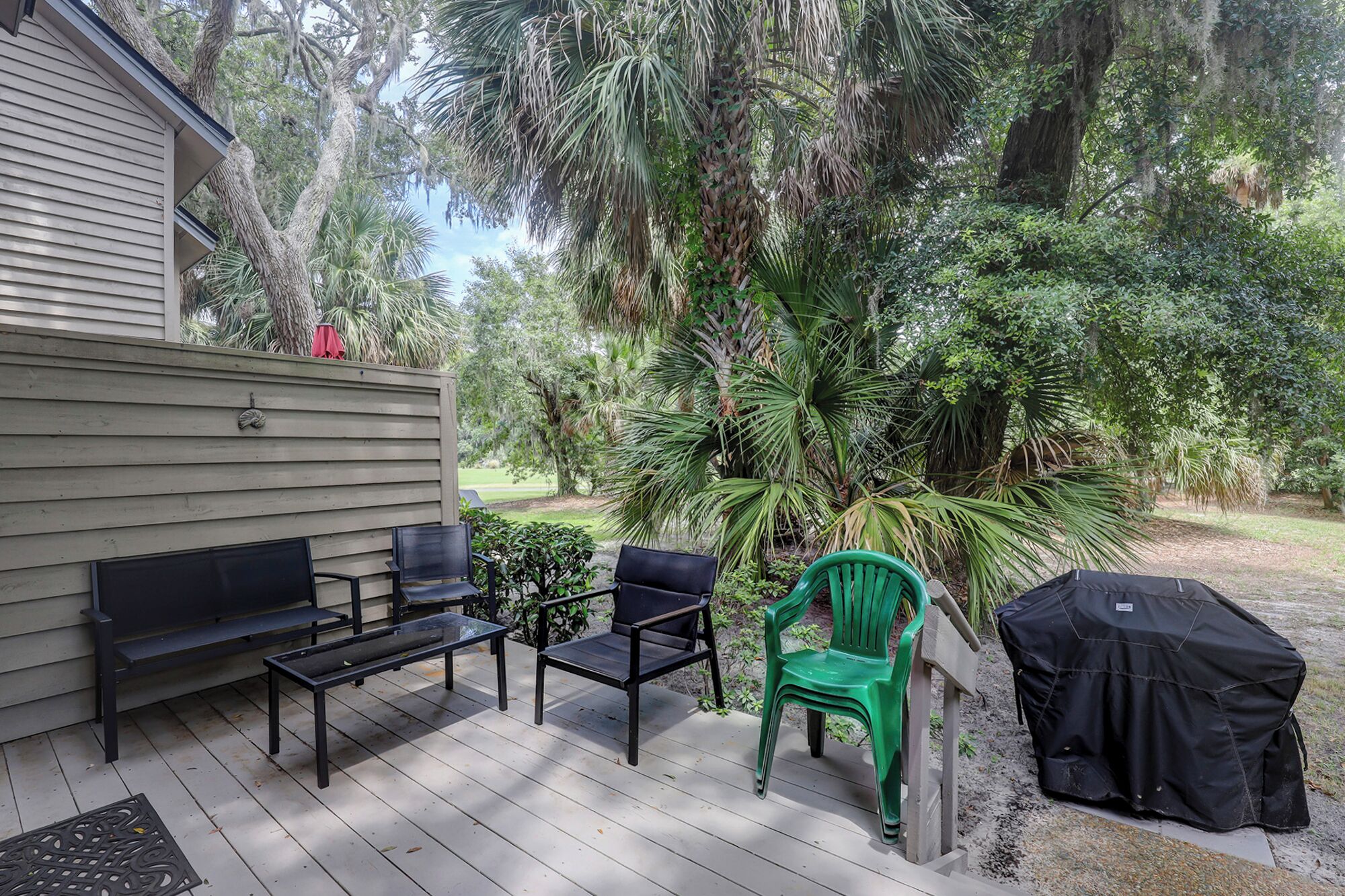 A covered patio with a wooden deck, two black chairs, a green chair, and a barbecue grill.
