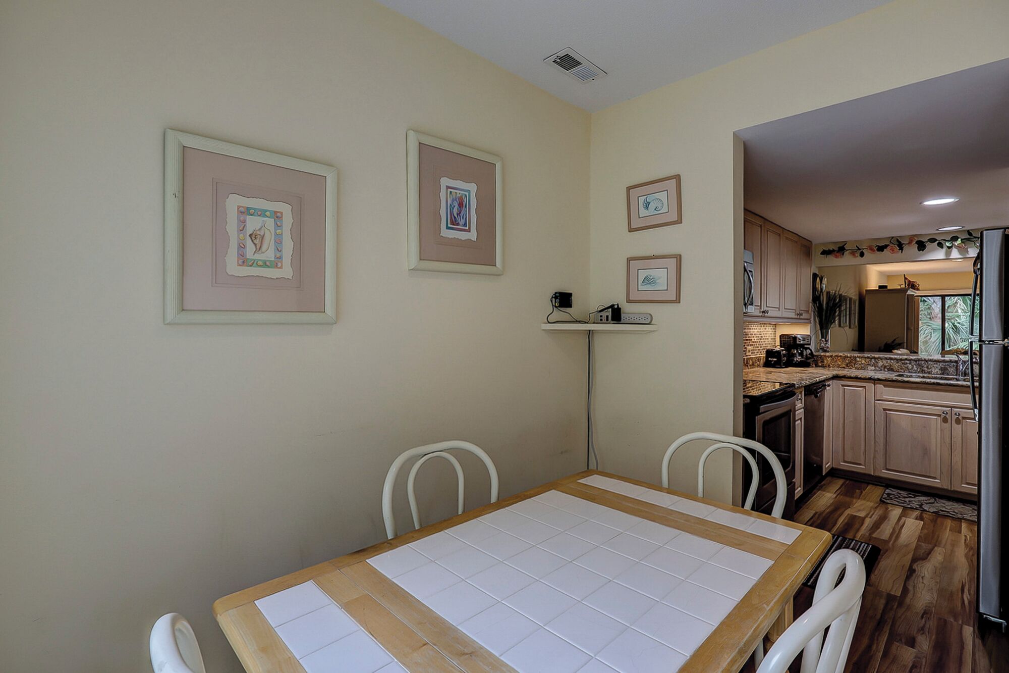 A dining area with a table and chairs, a kitchen in the background, and framed artwork on the wall.