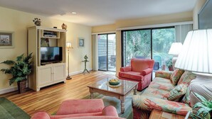 Una sala de estar con televisor, un sillón rojo, un sofá con cojines florales, una mesa de centro de vidrio y un ventanal corredero que da a un patio.