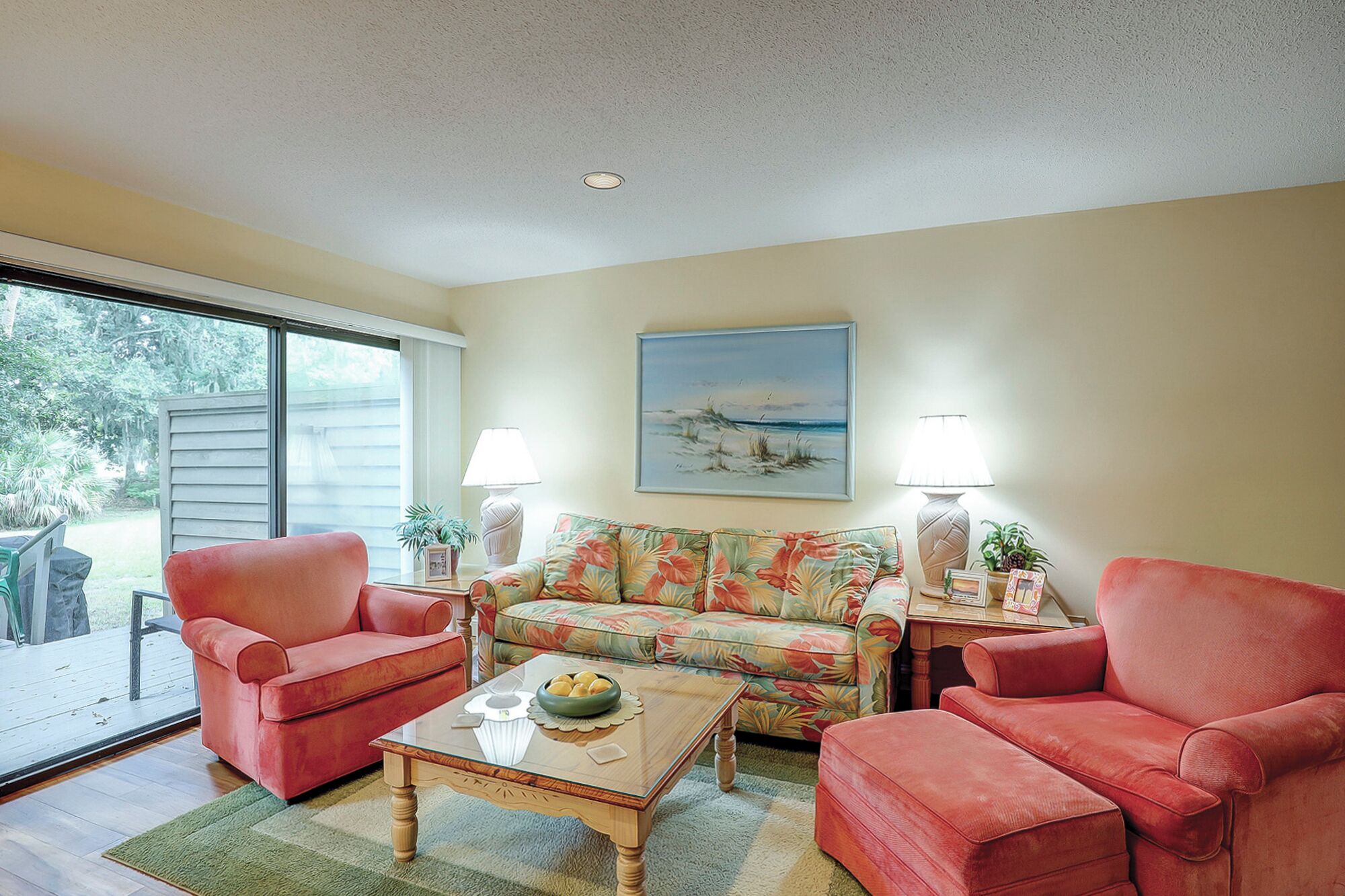 A living room with a glass coffee table, two red armchairs, a floral sofa, and two lamps.