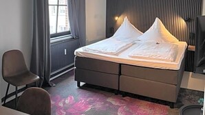 Quarto de hotel com cama, escrivaninha, cadeira, radiador e janela com cortinas.