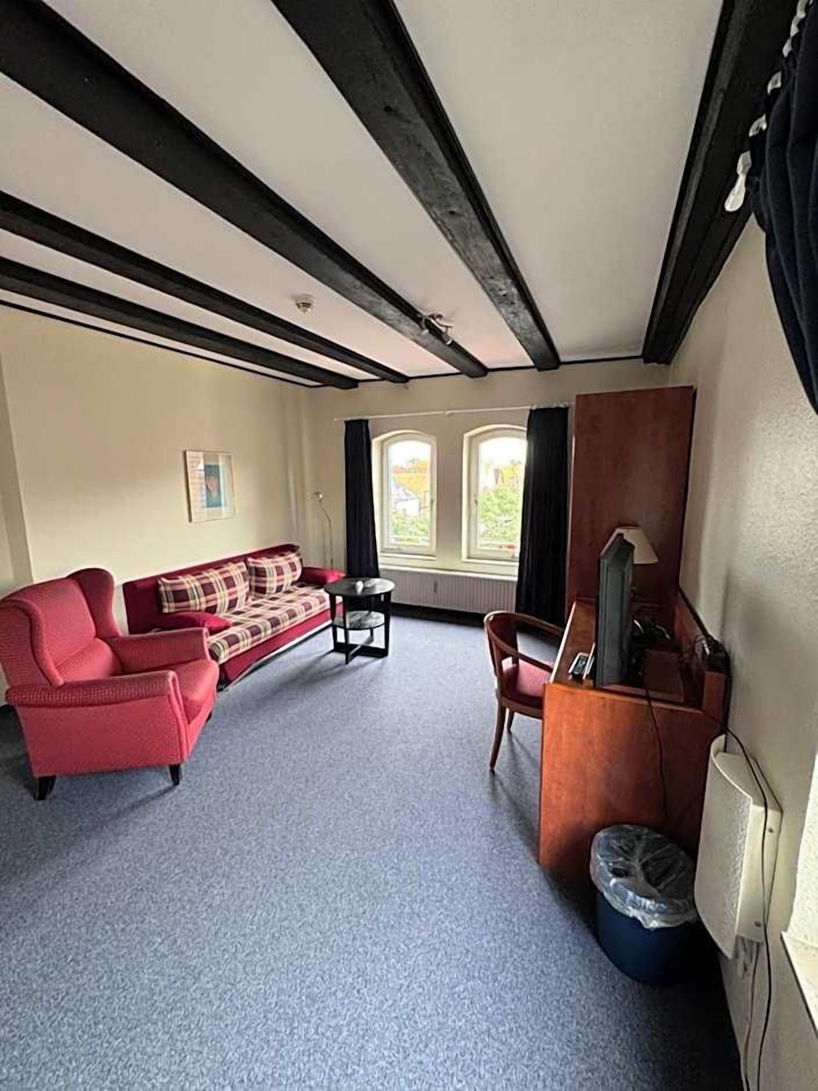 Ein Zimmer mit einem roten Sofa, einem kleinen Tisch, einer TV-Konsole und einem Fenster mit Vorhängen.