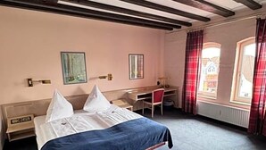 Quarto de hotel com cama, escrivaninha, cadeira e janela com cortinas.