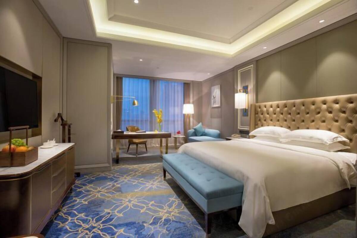 A hotel room with a large bed, a desk, a chair, a bench, and a TV.