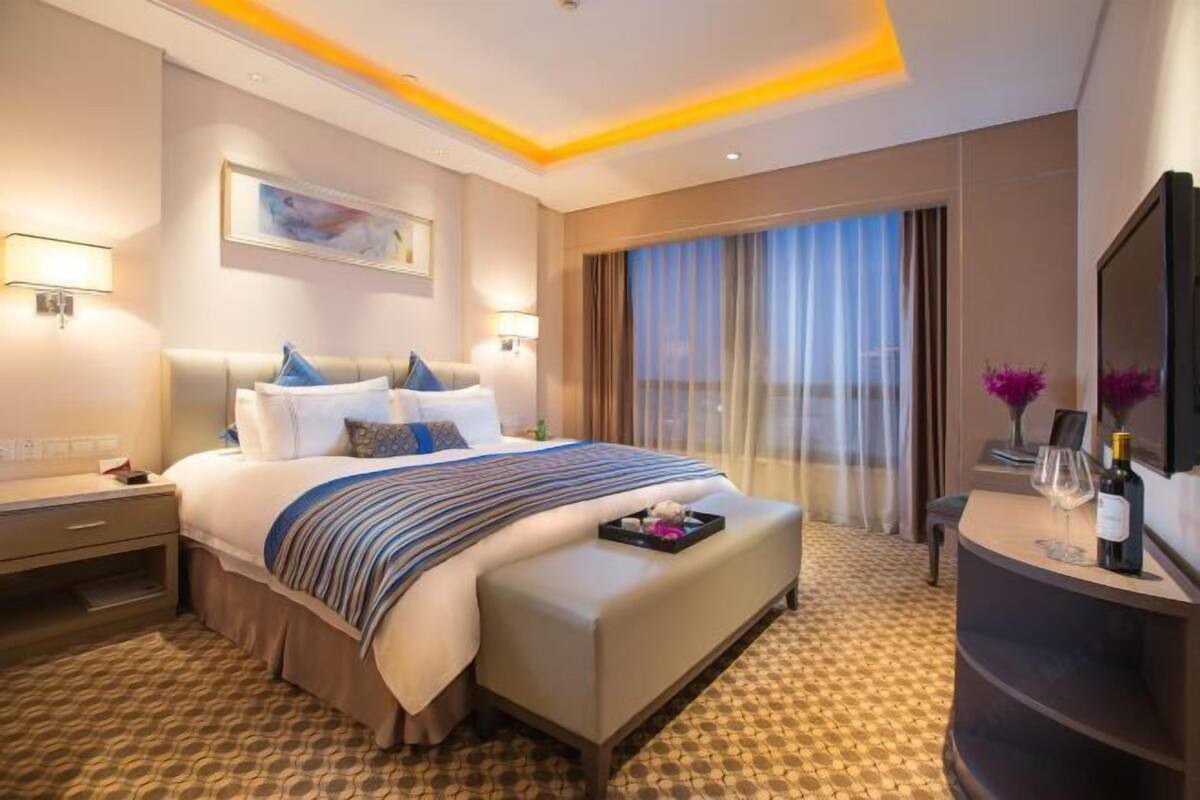 A hotel room with a large bed, a bedside table, a desk with a chair, a large window with curtains, and a wall-mounted TV.