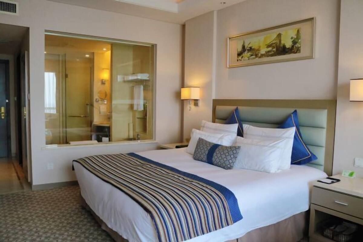 A hotel room with a large bed, a nightstand, a bedside table, a wall-mounted lamp, and a large window.