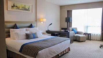 A hotel room with a large bed, a desk, a chair, and a view of the city through the window.