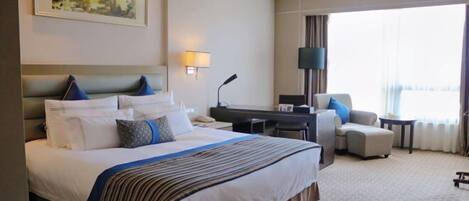 A hotel room with a large bed, a desk, a chair, and a view of the city through the window.