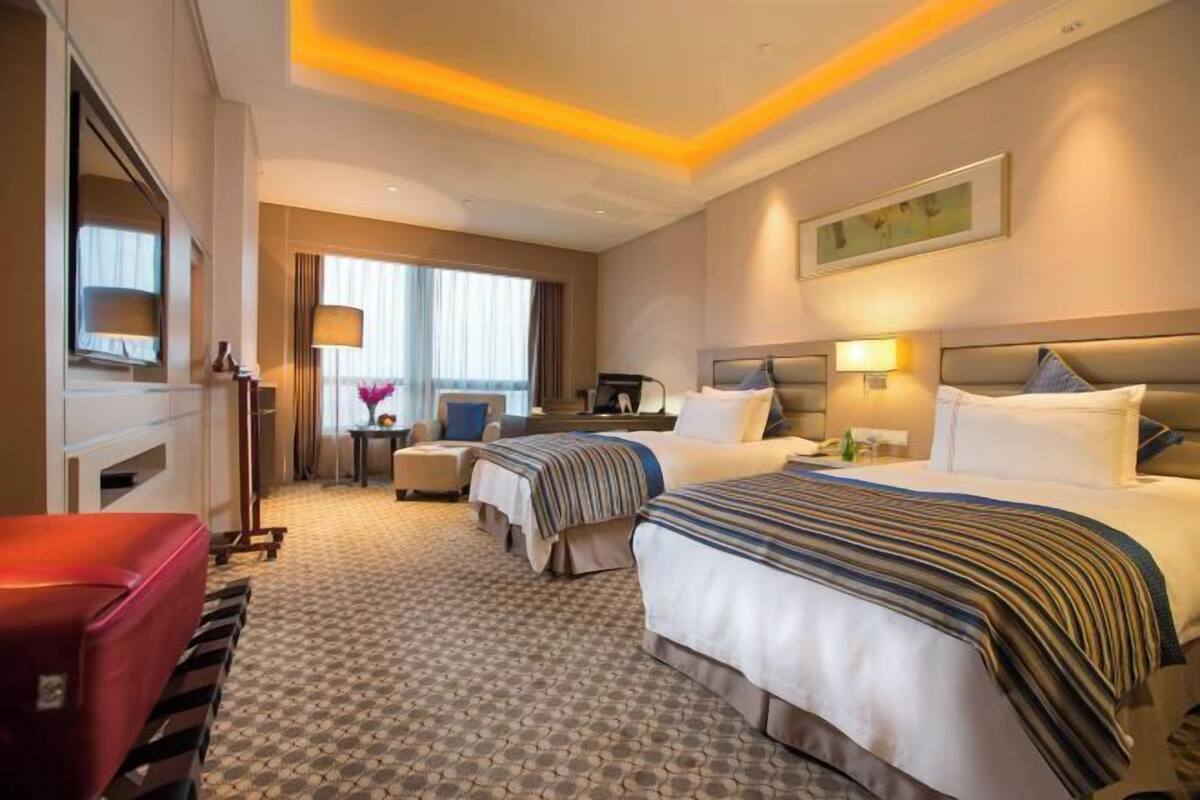 A hotel room with two beds, a desk, a chair, a red suitcase, and a window with curtains.