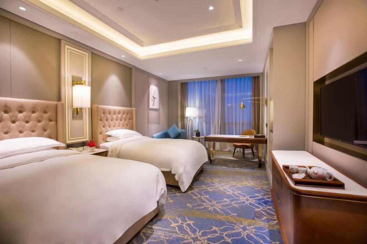 A hotel room with two beds, a desk, a TV, and a large window with curtains.