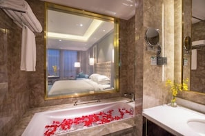 A modern bathroom with a large bathtub, a vanity with a mirror, and a view of a bedroom through a large mirror. - Hopesun Hotel (Jiangyin)