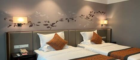 A hotel room with two beds, each with white bedding and orange patterned throw pillows. There are wall-mounted lamps on either side of the beds, and a small bedside table with a phone and a notepad.