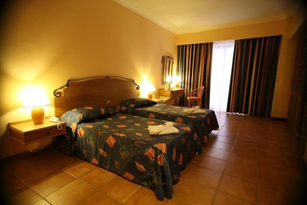 A hotel room with two beds, a nightstand with a lamp, a chair, and a window with curtains.