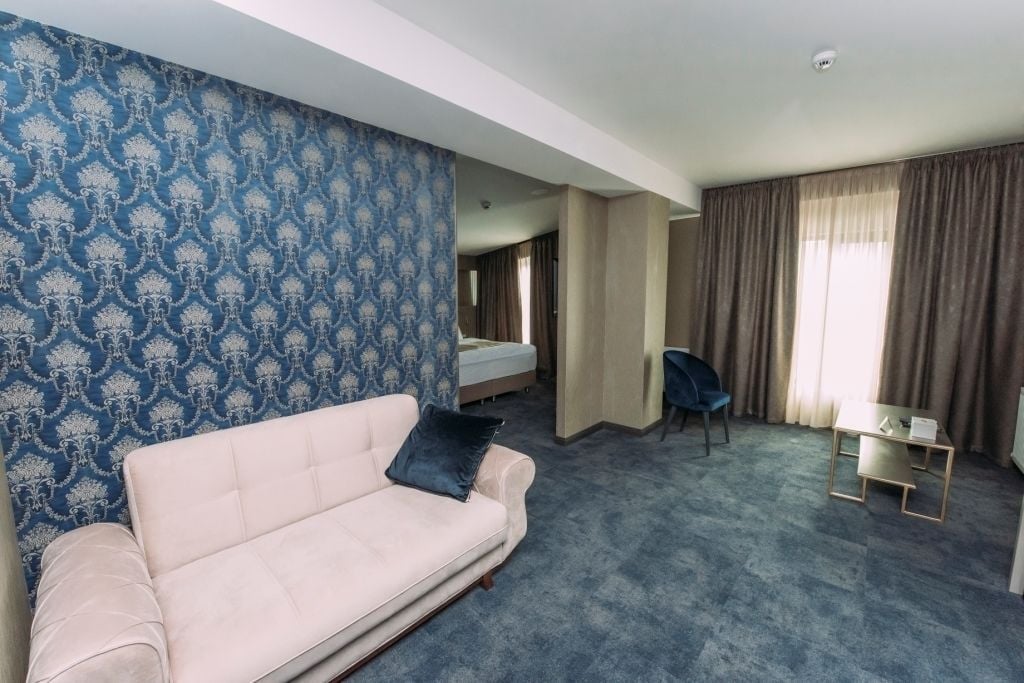A hotel room with a patterned blue wall, a light-colored sofa, a small table, a chair, and a bed in the background.