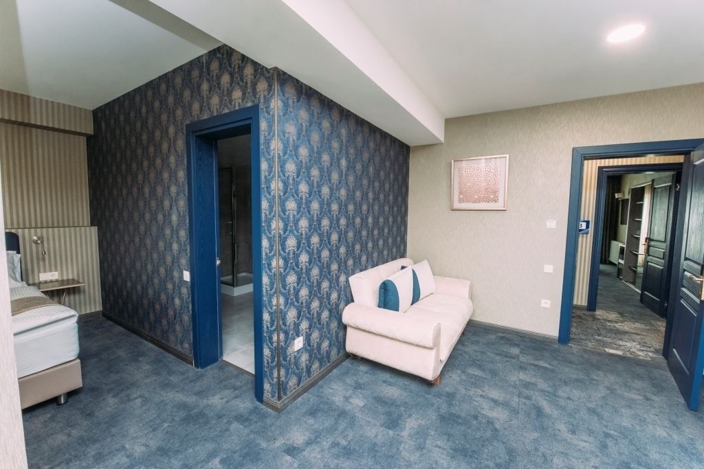 Une pièce avec un papier peint bleu et blanc à motifs, un canapé blanc agrémenté de coussins bleus, un lit recouvert d’une couverture à rayures et un tableau encadré accroché au mur.