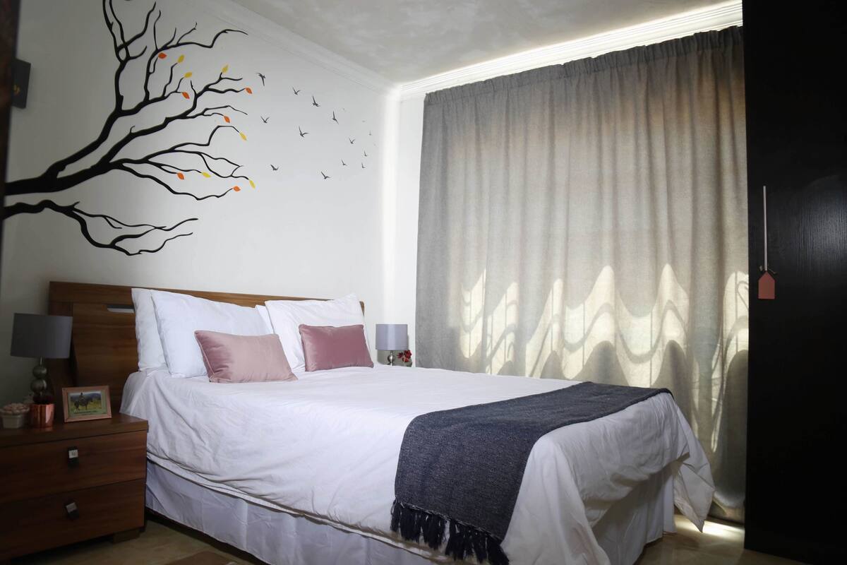 A neatly made bed with white linens and a dark throw blanket, a wooden nightstand with a lamp, a wall-mounted tree decal, and a large window with sheer curtains.