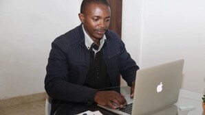 A person using a laptop at a table with a white wall and a wooden door in the background. - Denver Echo- Executive Guest House (Maseru)