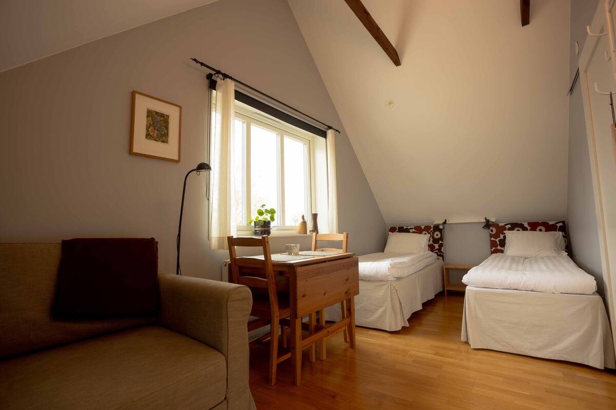 Ein gemütliches, helles Zimmer mit einem Holztisch, zwei Einzelbetten, einer Couch und einem Fenster mit Vorhängen.