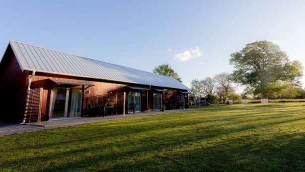 A modern building with a sloped metal roof, large glass doors, and a covered outdoor seating area. - Ljungs Fem Rum (Gotlands Tofta)