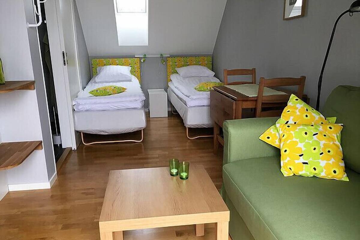 Ein kleiner, gut beleuchteter Raum mit zwei Einzelbetten, einem grünen Sofa, einem Holztisch und einem Fenster, das natürliches Licht hereinlässt.