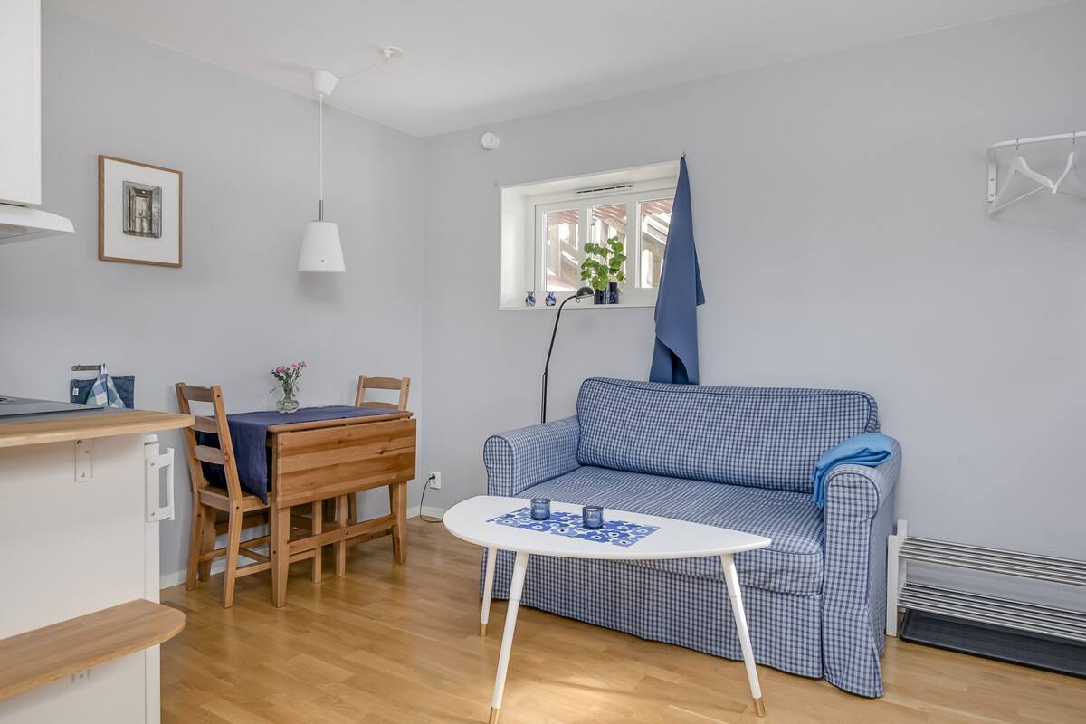 Ein kompakter Wohnbereich mit einem karierten Sofa, einem kleinen Esstisch, einer weißen Pendelleuchte und einem Fenster mit einer Pflanze.