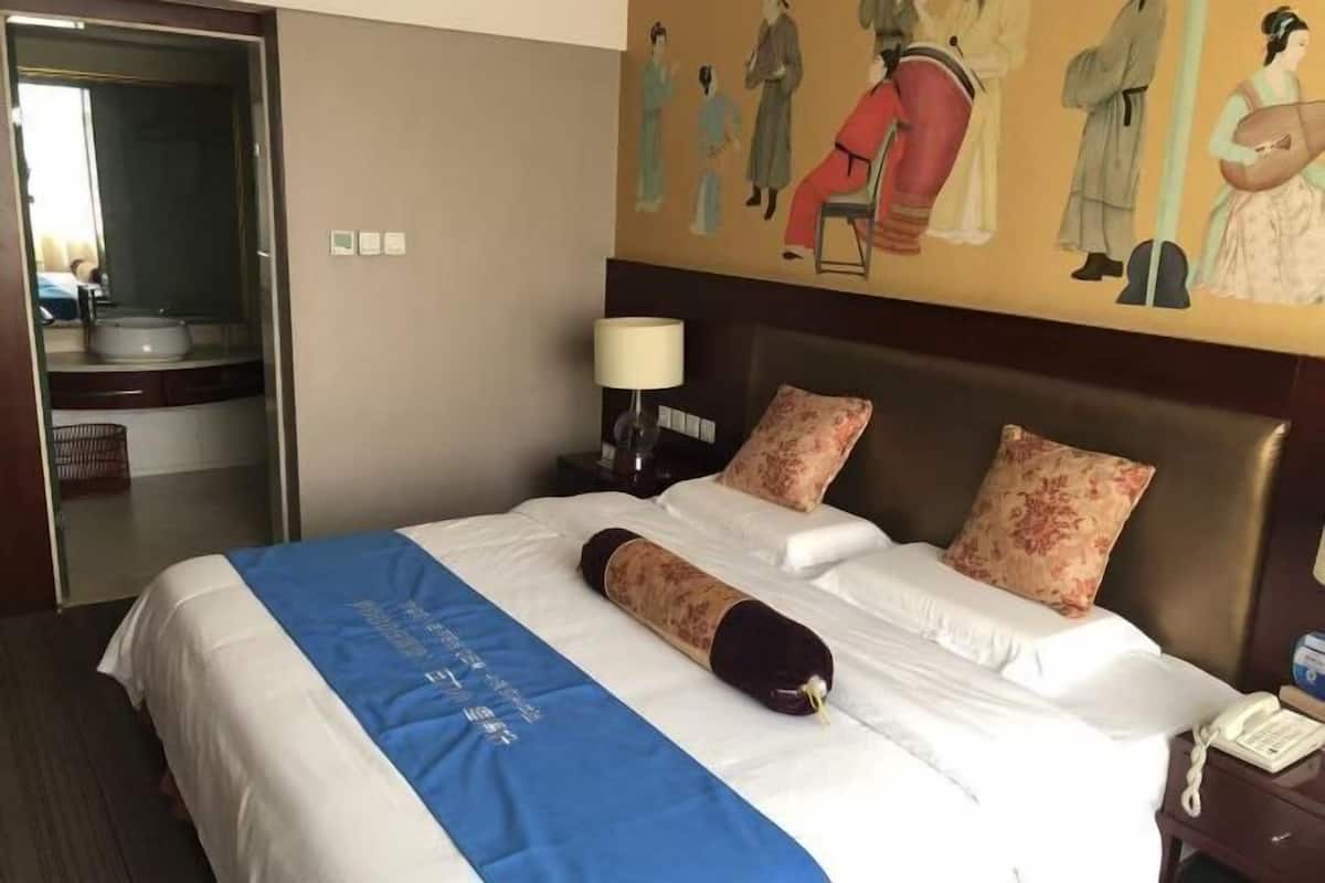 Une chambre d’hôtel avec un lit, une table de chevet avec une lampe, un tableau au mur et une salle de bain visible à travers une porte ouverte.