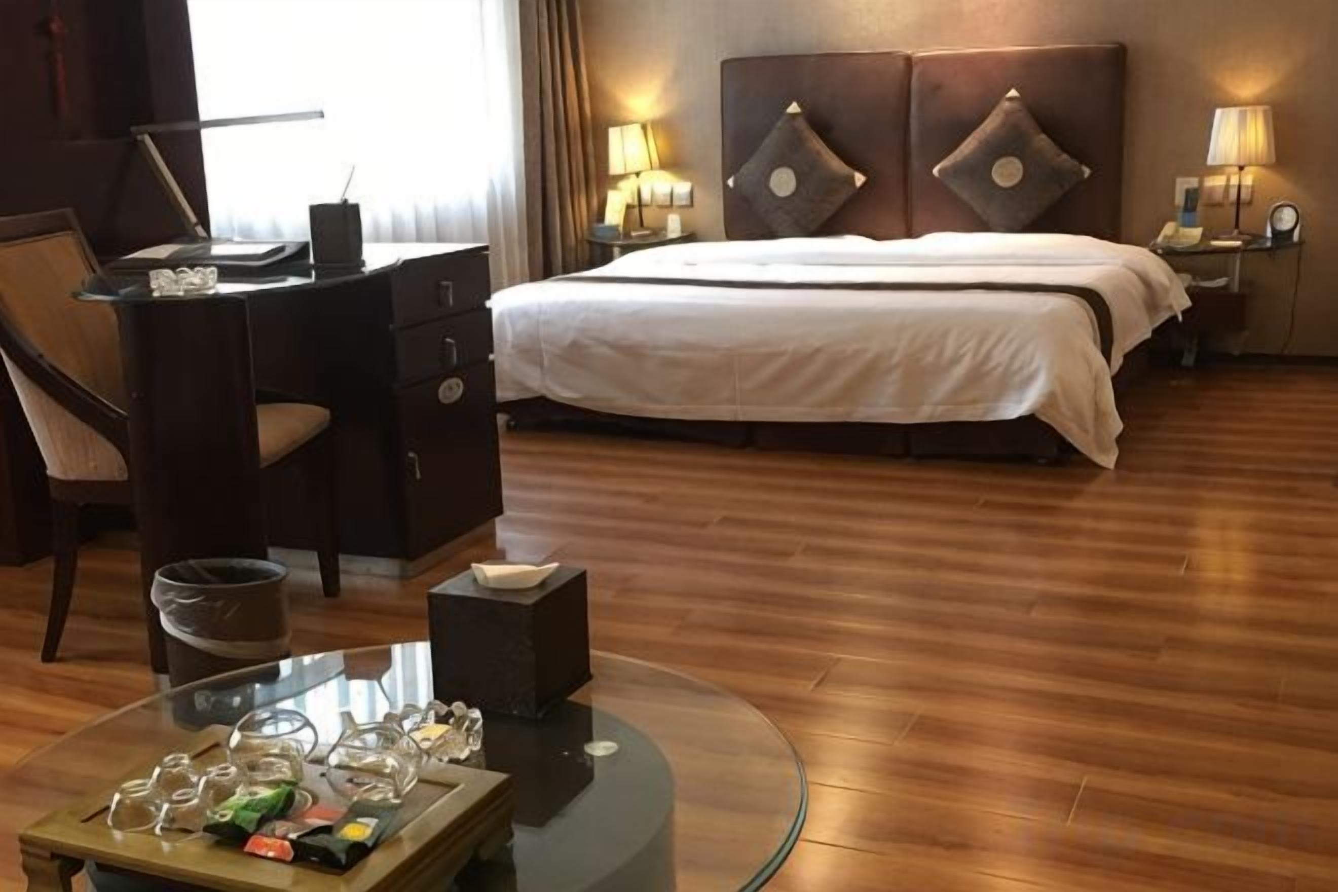 A hotel room with a bed, a desk, a chair, a glass coffee table, and a nightstand with lamps.