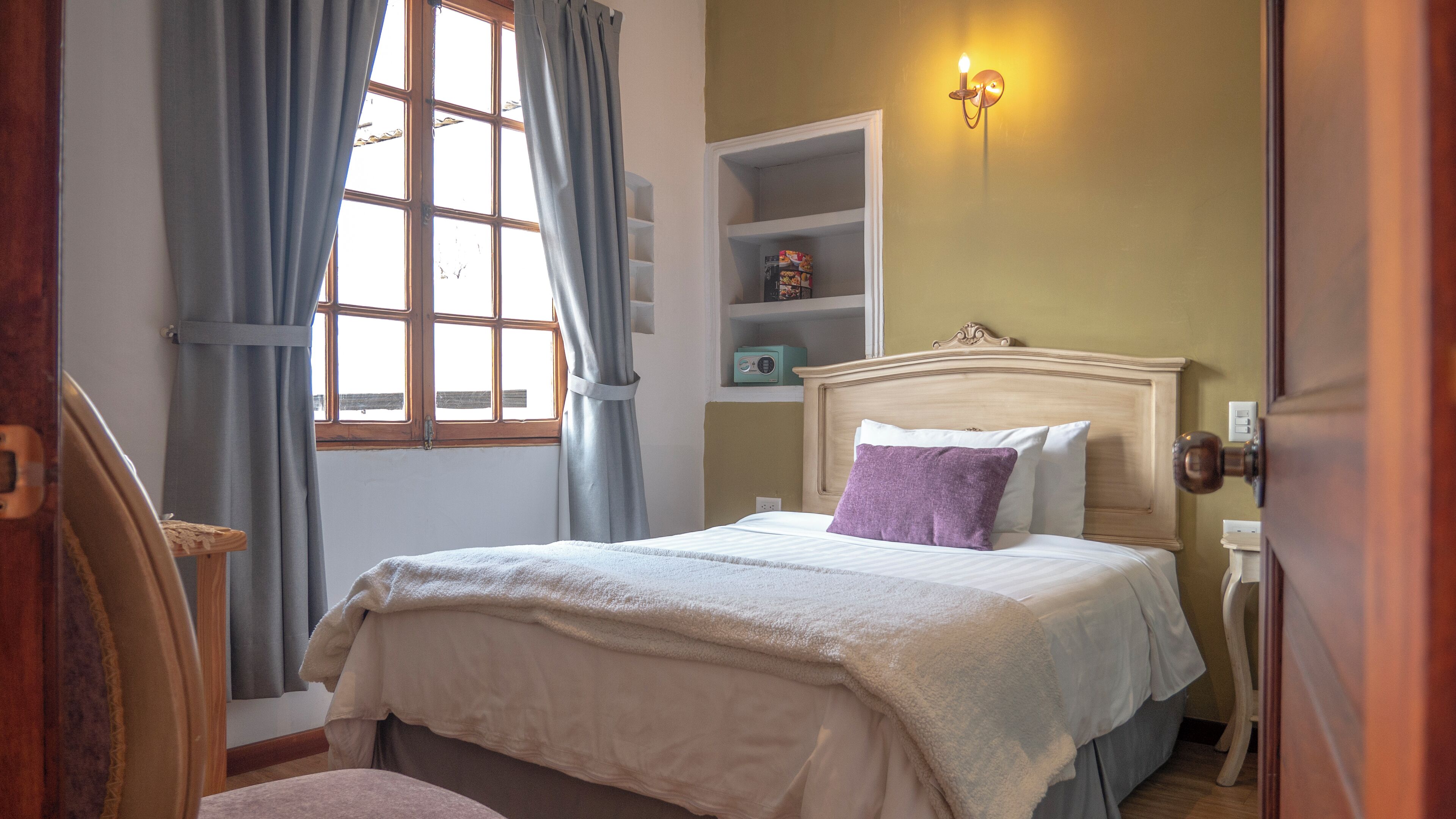 Un dormitorio con cama, mesita de noche, estante con radio y ventana con cortinas.